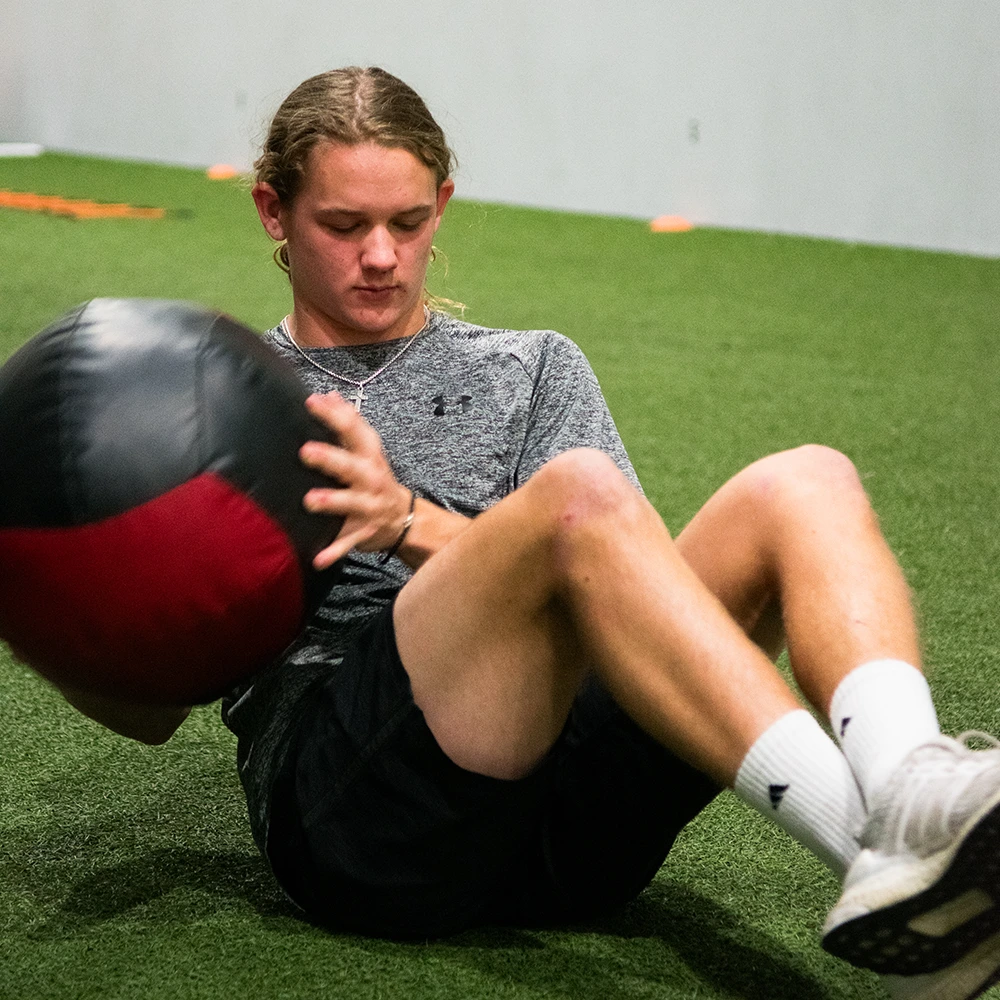 Teen male sitting on turf in a gym doing med ball russian twists.