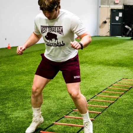 A young man wearing a West Texas A & M shirt while exercising on a agility ladder inside an athletic gym with turf.