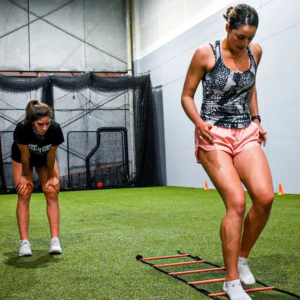Two teen girls exercise on an agility ladder inside an athletic gym with turf.