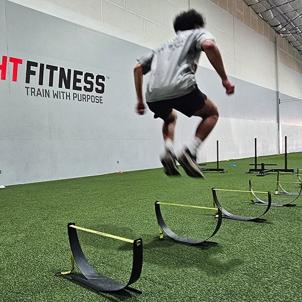Teen boy jumping over mini hurdles inside a gym with turf.